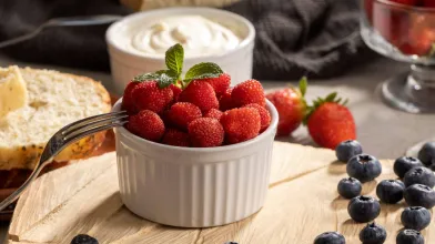 A small bowl filled with berries sits on a wooden plate, with blueberries scattered around it and other fruit and cream in the background.