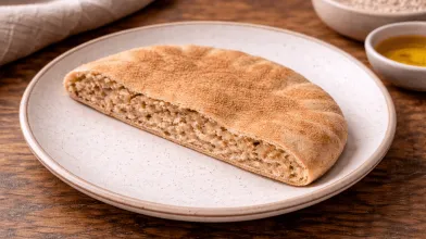 Whole-grain flatbread on a plate, showing its textured interior. Grains and olive oil nearby.