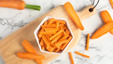 Bowl of carrot sticks on a cutting board with whole carrots and more sticks on marble.