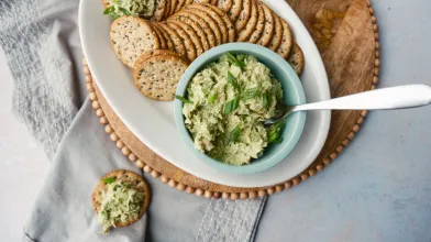 Whole-grain crackers with tuna avocado spread in a blue bowl and on crackers, garnished with herbs.