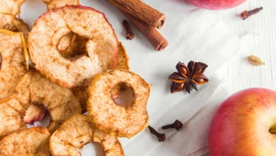 Dried apple slices, whole red apples, cinnamon sticks, and spices on a white surface.