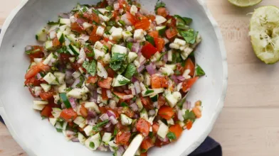 Fresh salsa with diced tomatoes, zucchini, onions, and herbs in a white bowl, with lime.