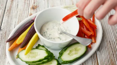 Hand dips red bell pepper into herb dip, surrounded by colorful veggies.