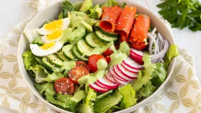 Salad bowl with lettuce, cucumber, tomato, egg, radish, salmon, avocado, and green dressing.