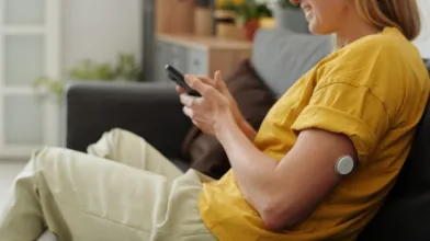 Smiling woman on a couch uses phone, a continuous glucose monitor visible on her upper arm.