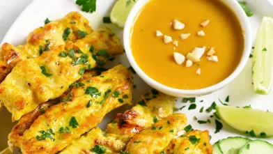 Chicken satay skewers, peanut sauce, lime wedges, and cucumber slices on a plate.