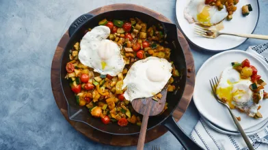 Skillet with colorful vegetable hash, two fried eggs. Two servings ready on plates.