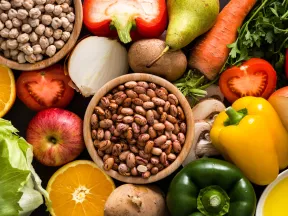 Overhead shot of bowl of chickpeas, bowl of beans, an apple, an orange, mushrooms, bell peppers, pears, kiwis, carrots, tomato, onion, and herbs.