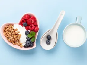 Heart-shaped bowl with yogurt, granola, raspberries, blueberries; spoons; mug of milk.