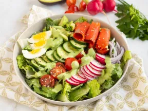 Salad bowl with lettuce, cucumber, tomato, egg, radish, salmon, avocado, and green dressing.