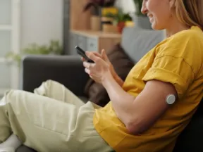 Smiling woman on a couch uses phone, a continuous glucose monitor visible on her upper arm.