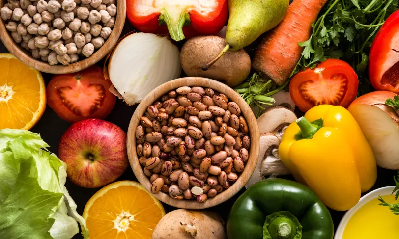 Overhead shot of bowl of chickpeas, bowl of beans, an apple, an orange, mushrooms, bell peppers, pears, kiwis, carrots, tomato, onion, and herbs.