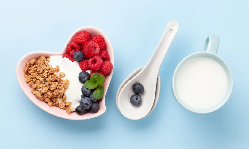 Heart-shaped bowl with yogurt, granola, raspberries, blueberries; spoons; mug of milk.