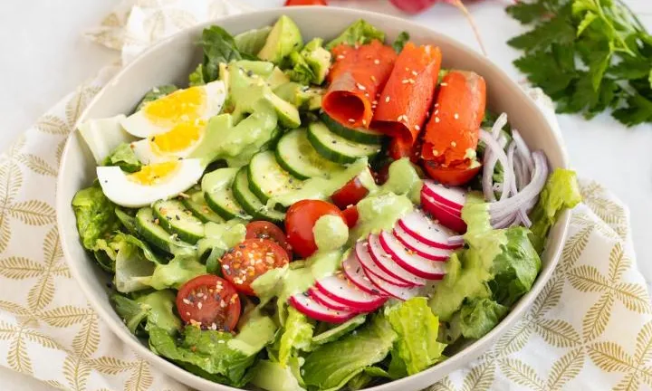 Salad bowl with lettuce, cucumber, tomato, egg, radish, salmon, avocado, and green dressing.