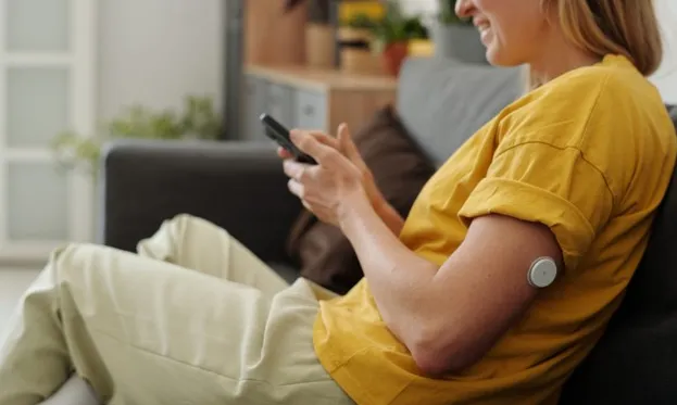 Smiling woman on a couch uses phone, a continuous glucose monitor visible on her upper arm.