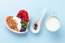 Heart-shaped bowl with yogurt, granola, raspberries, blueberries; spoons; mug of milk.