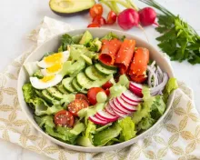 Salad bowl with lettuce, cucumber, tomato, egg, radish, salmon, avocado, and green dressing.