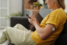 Smiling woman on a couch uses phone, a continuous glucose monitor visible on her upper arm.