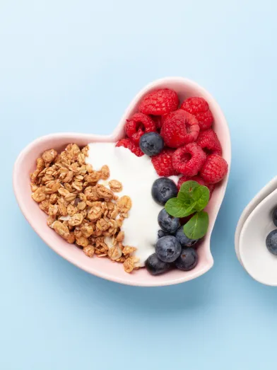 Heart-shaped bowl with yogurt, granola, raspberries, blueberries; spoons; mug of milk.