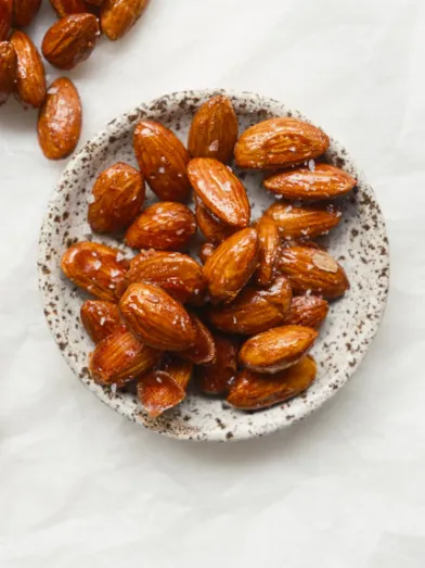 Glazed almonds in a small bowl, with more scattered on white.