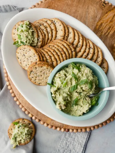 Whole-grain crackers with tuna avocado spread in a blue bowl and on crackers, garnished with herbs.
