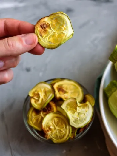 Hand holds a baked zucchini chip; bowl of chips and fresh zucchini.
