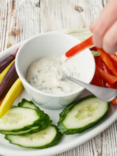 Hand dips red bell pepper into herb dip, surrounded by colorful veggies.