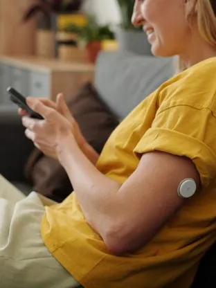 Smiling woman on a couch uses phone, a continuous glucose monitor visible on her upper arm.
