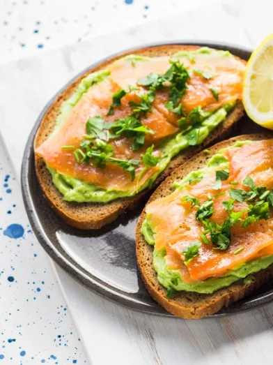 Two slices of toast topped with mashed avocado, smoked salmon, and herbs