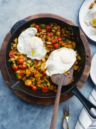 Skillet with colorful vegetable hash, two fried eggs. Two servings ready on plates.