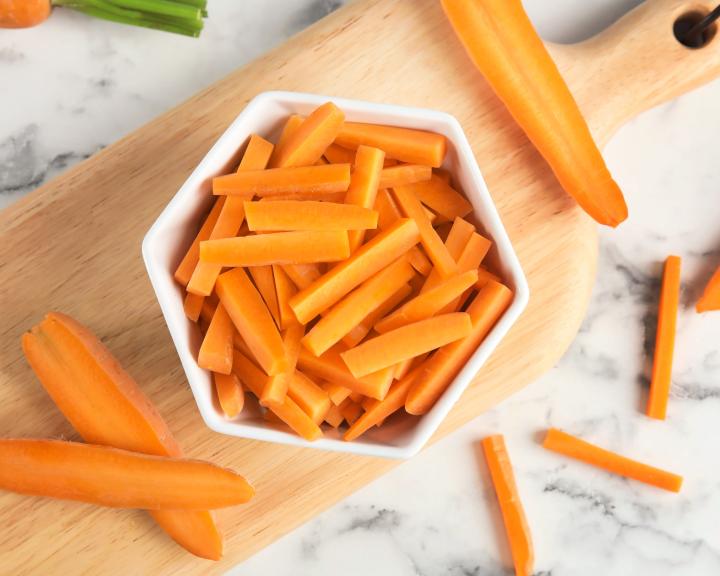 Bowl of carrot sticks on a cutting board with whole carrots and more sticks on marble.
