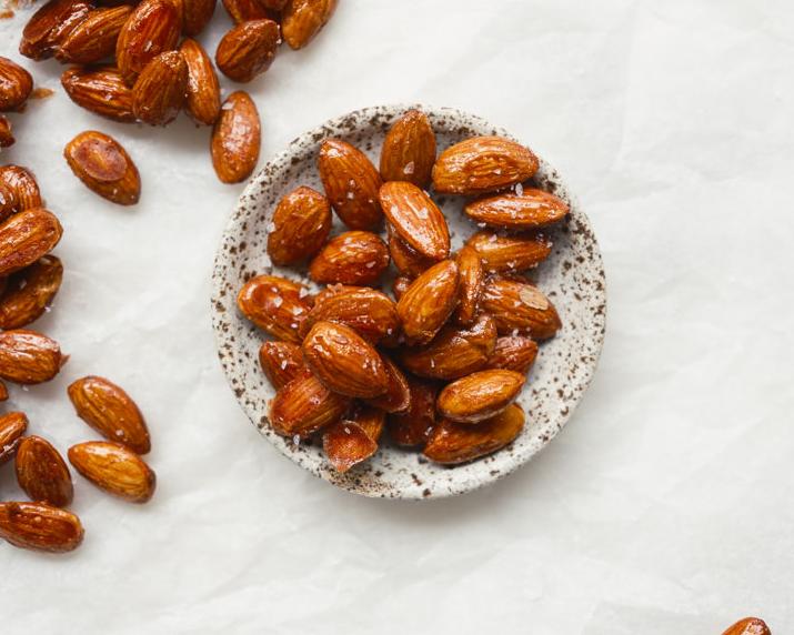 Glazed almonds in a small bowl, with more scattered on white.