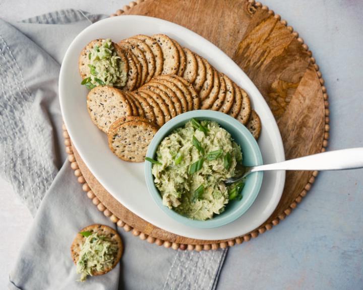 Whole-grain crackers with tuna avocado spread in a blue bowl and on crackers, garnished with herbs.