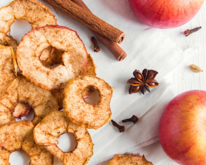 Dried apple slices, whole red apples, cinnamon sticks, and spices on a white surface.