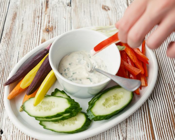 Hand dips red bell pepper into herb dip, surrounded by colorful veggies.