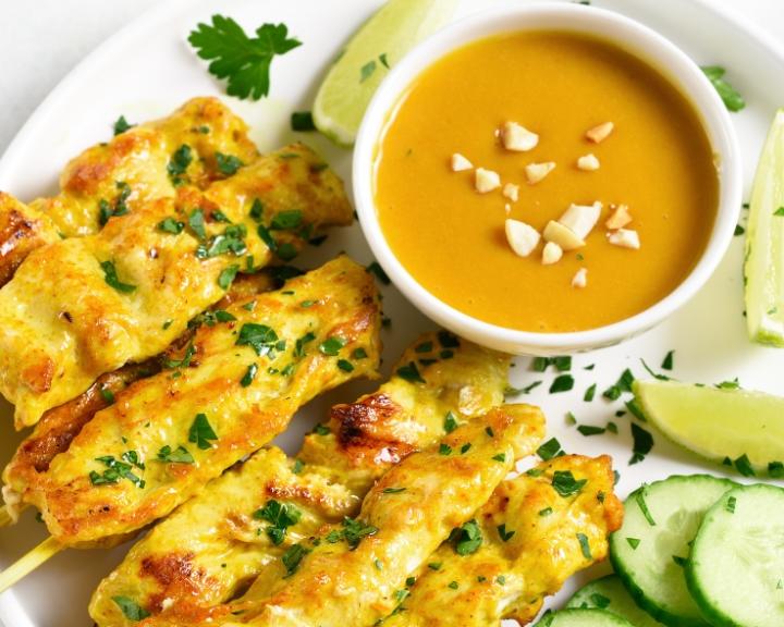 Chicken satay skewers, peanut sauce, lime wedges, and cucumber slices on a plate.
