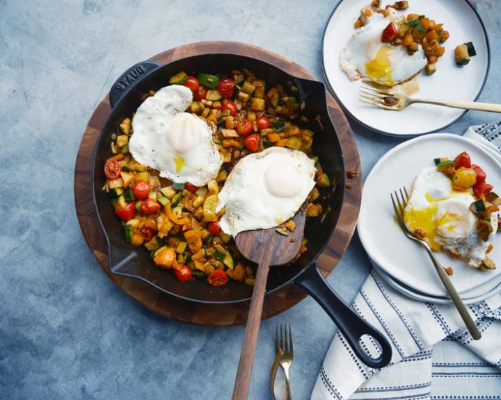 Skillet with colorful vegetable hash, two fried eggs. Two servings ready on plates.