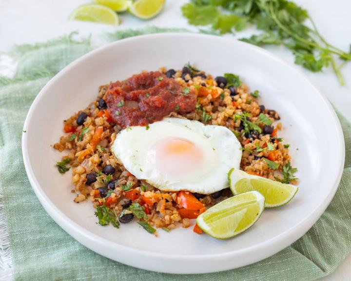 A bowl with cauliflower rice, peppers and black beans topped with a fried egg. Served with lime wedges. 